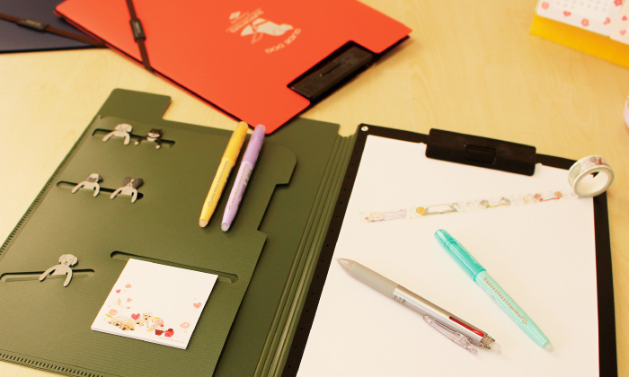 オリーブのバインダーが机の上に置かれている。バインダー左側のポケットには。マーカーペンや紙クリップ。付箋が挟んである。バインダー右側には紙が挟んであり。マスキングテープを貼っている。紙の上にマーカーペンと2色ボールペン。