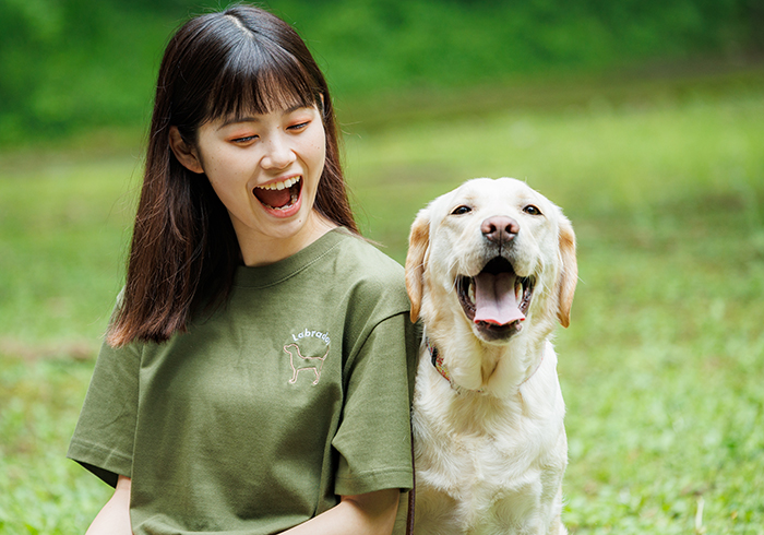 女性がラブラドール刺しゅうのカーキのTシャツを着て、隣に座るラブラドールと同じ笑顔の写真