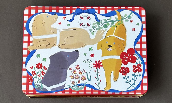 赤のギンガムチェックの背景にブルーリボンが一周その中にゴールデン、イエローラブ、ブラックラブの盲導犬が描かれ、その周りには赤の可憐な花と葉のグリーンが描かれている