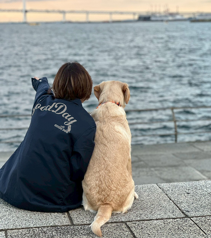 ネイビーのジャケットを着てラブラドールと海辺に腰掛け二人で海を見ている写真