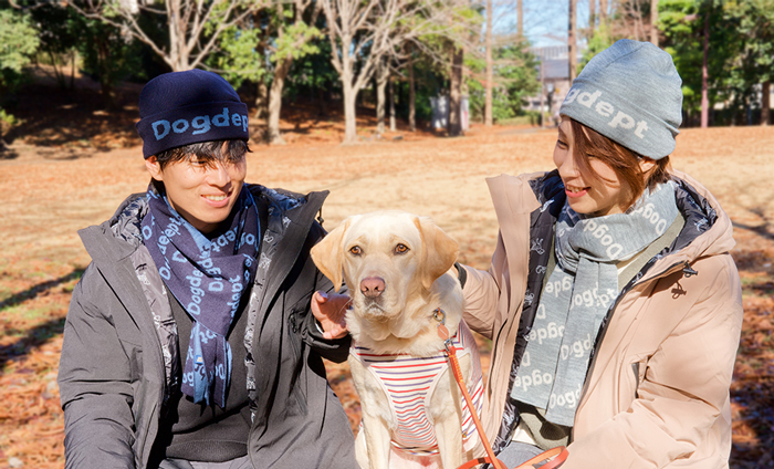 男性がネイビーのニットキャップとマフラーをし、女性がミックスグレーのニットキャップとマフラーを着けて真ん中にラブラドールといる写真