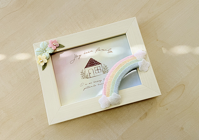 テーブルにフォトフレームが平置きして日が差し、虹と花のちりめん細工が立体的に見えている写真