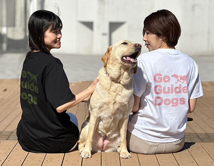 ２人の女性がブラックにオリーブのプリントのTシャツとホワイトにダスティピンクのプリントのTシャツを着ていて、間にラブラドールが座っている写真