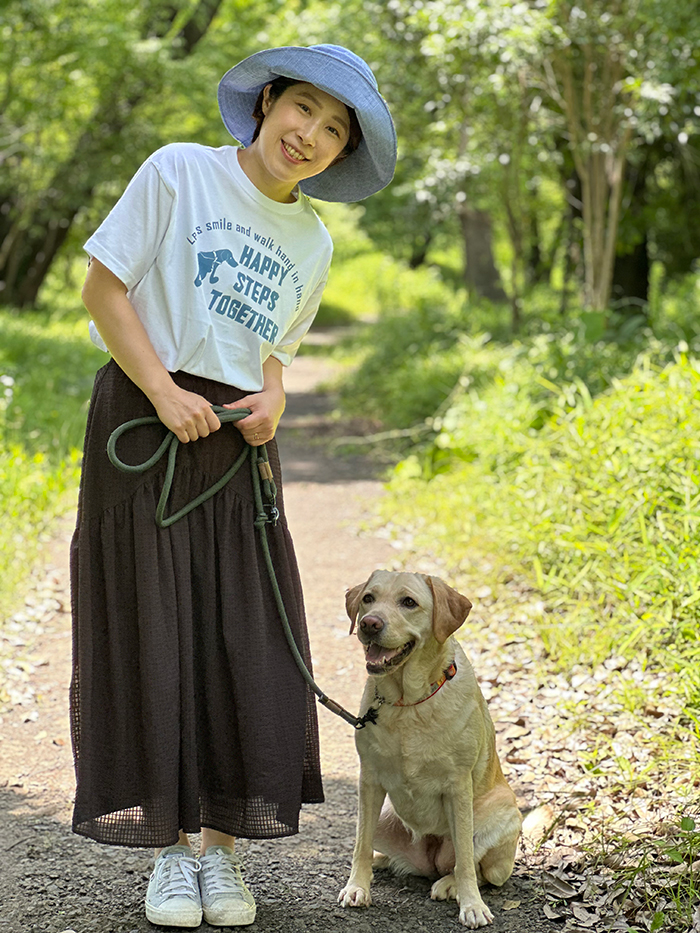 女性がホワイトのTシャツの下に黒いスカートを着用している写真。となりにはラブラドールが座っている。