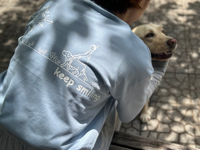 女性がアシットブルーのTシャツを着用しラブラドールを抱きしめて、背中のプリントが見えている写真。